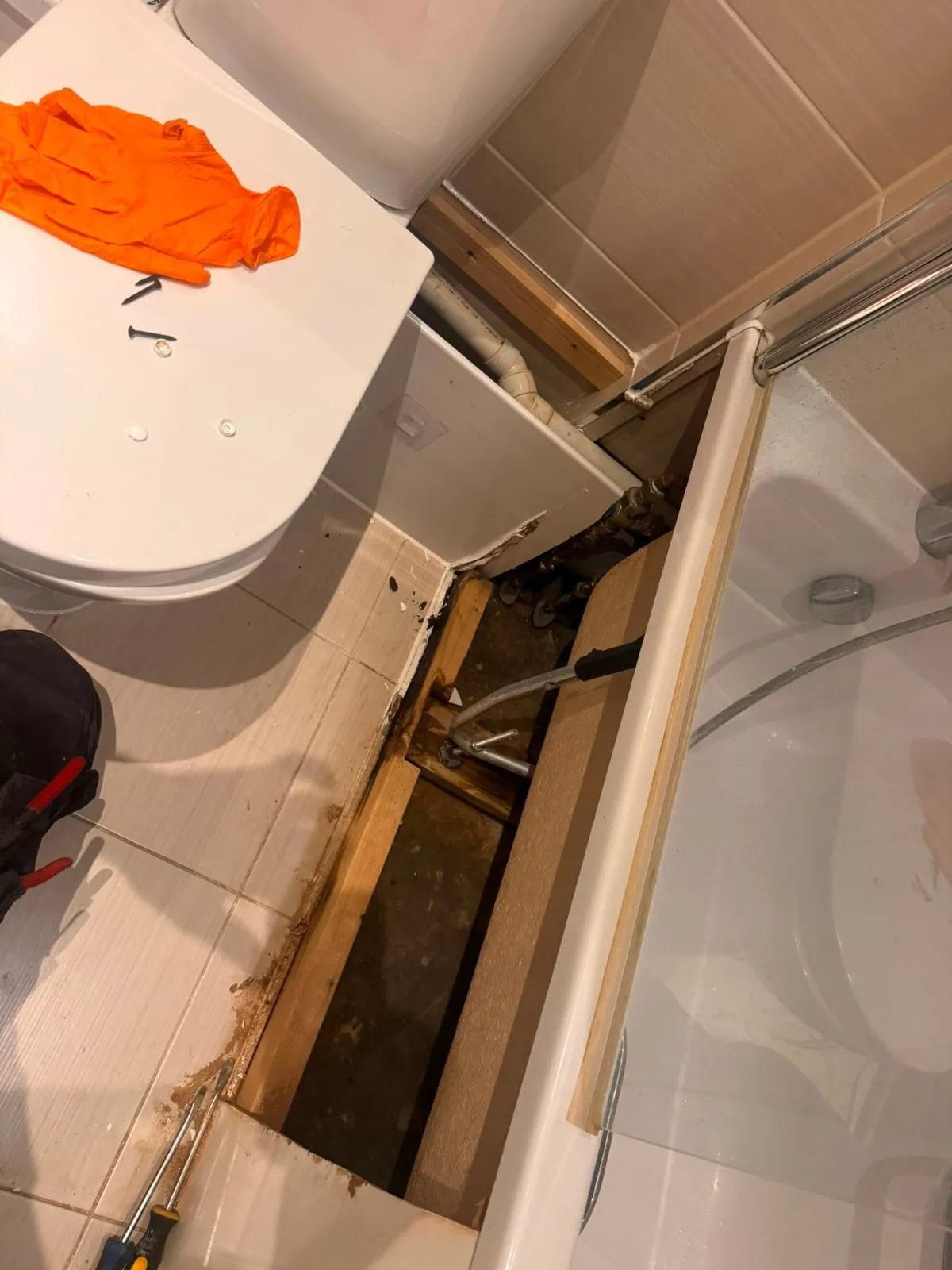 Bathroom with the bath side panel removed to show a dark floor void beside the toilet, with an orange glove on the lid