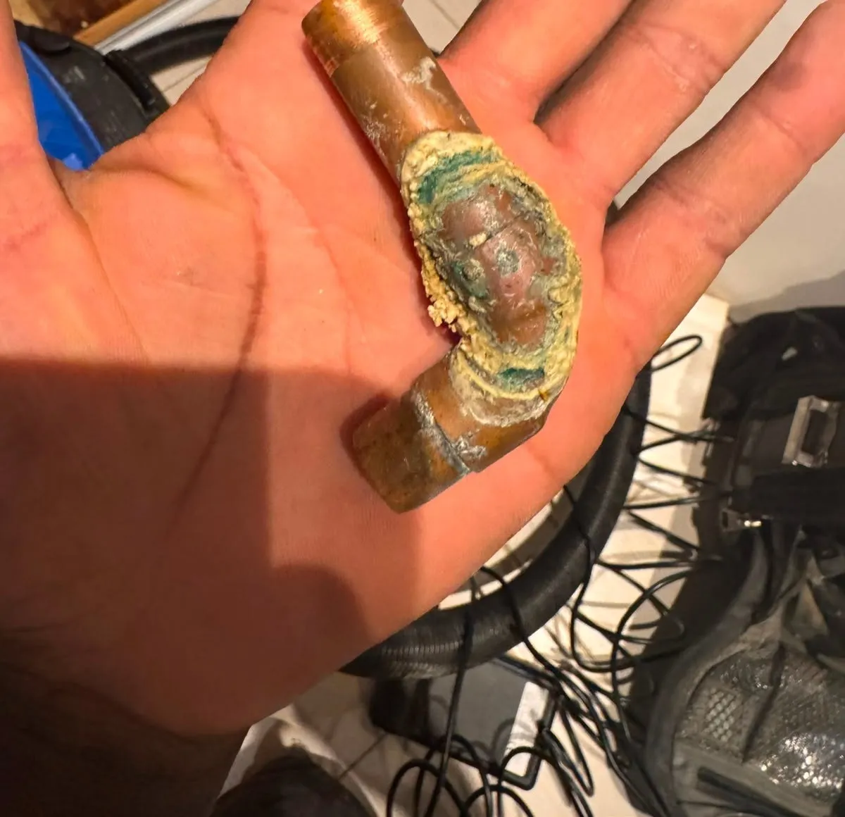 Short cut-out piece of copper pipe with an elbow, held in a palm, with green and cream crust around the bend
