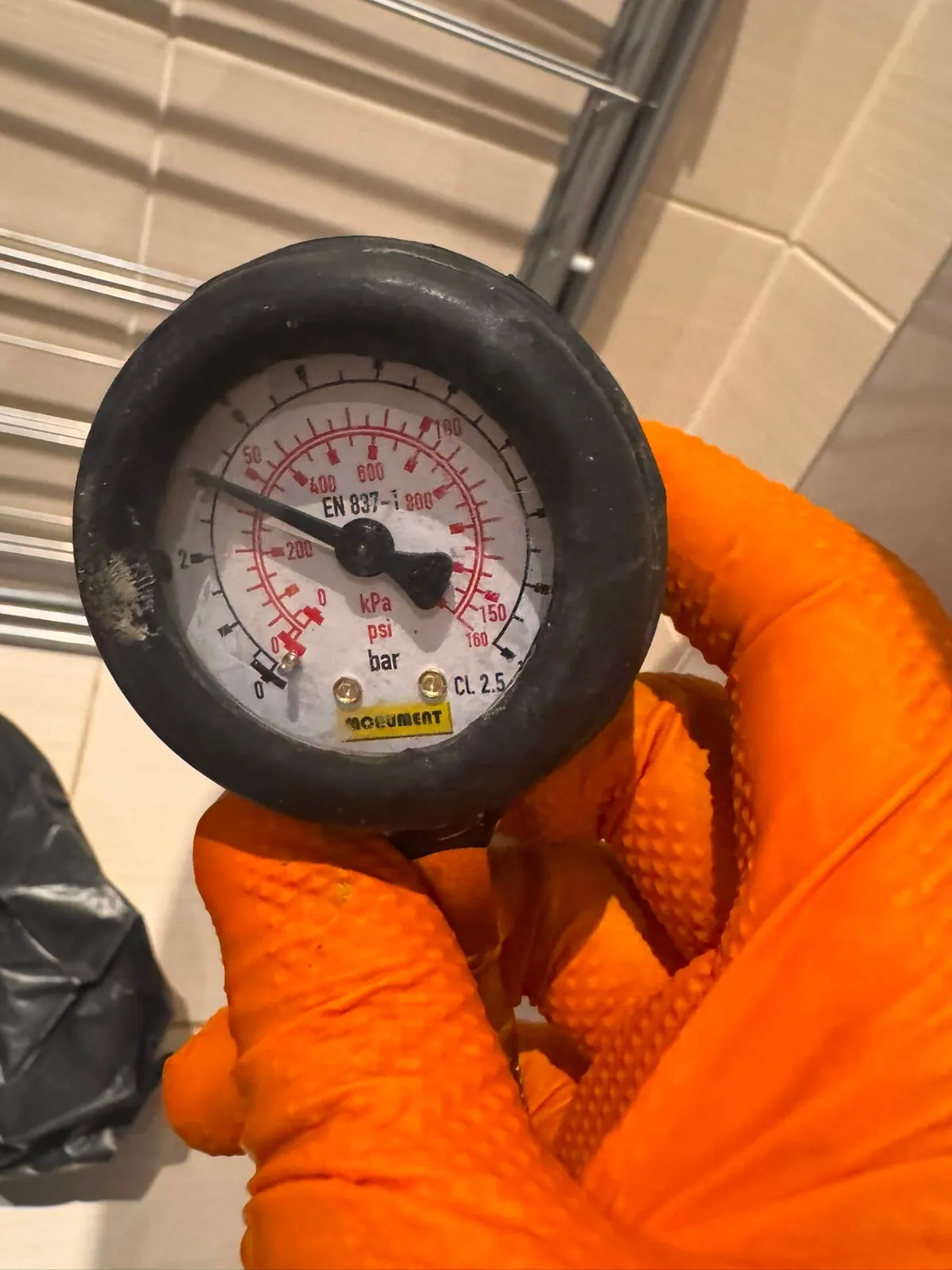 Pressure gauge with bar, kPa and psi scales held in an orange glove against a tiled bathroom wall