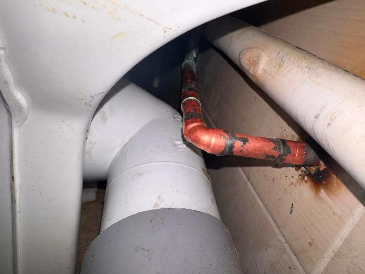 Copper supply pipe with an elbow and black patches, running beside a white toilet pan and a grey waste connector