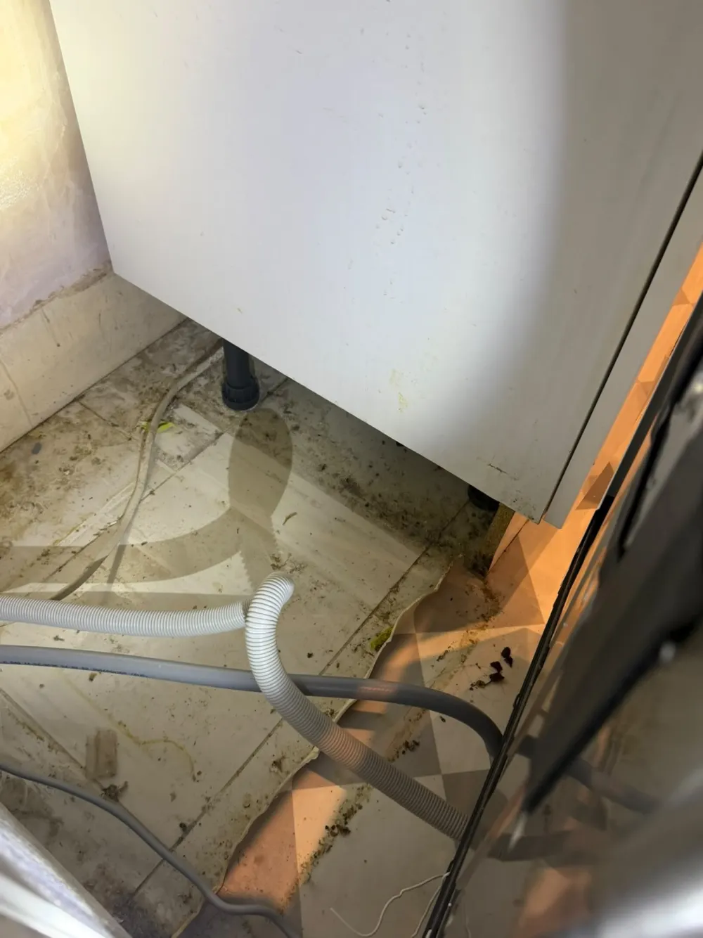 Ribbed grey hose and a smooth grey hose lying across a dirty tiled floor beside a white kitchen cabinet and dishwasher