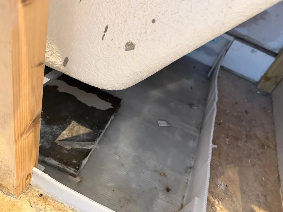 Underside of a white bath above a bare floor void, with a timber batten at the left and a stained board below it