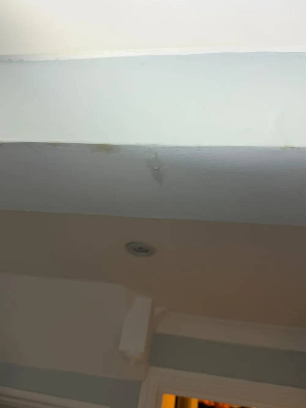 Pale water stain and a flaking paint patch on a blue-grey wall just below a white ceiling edge, with a recessed light below