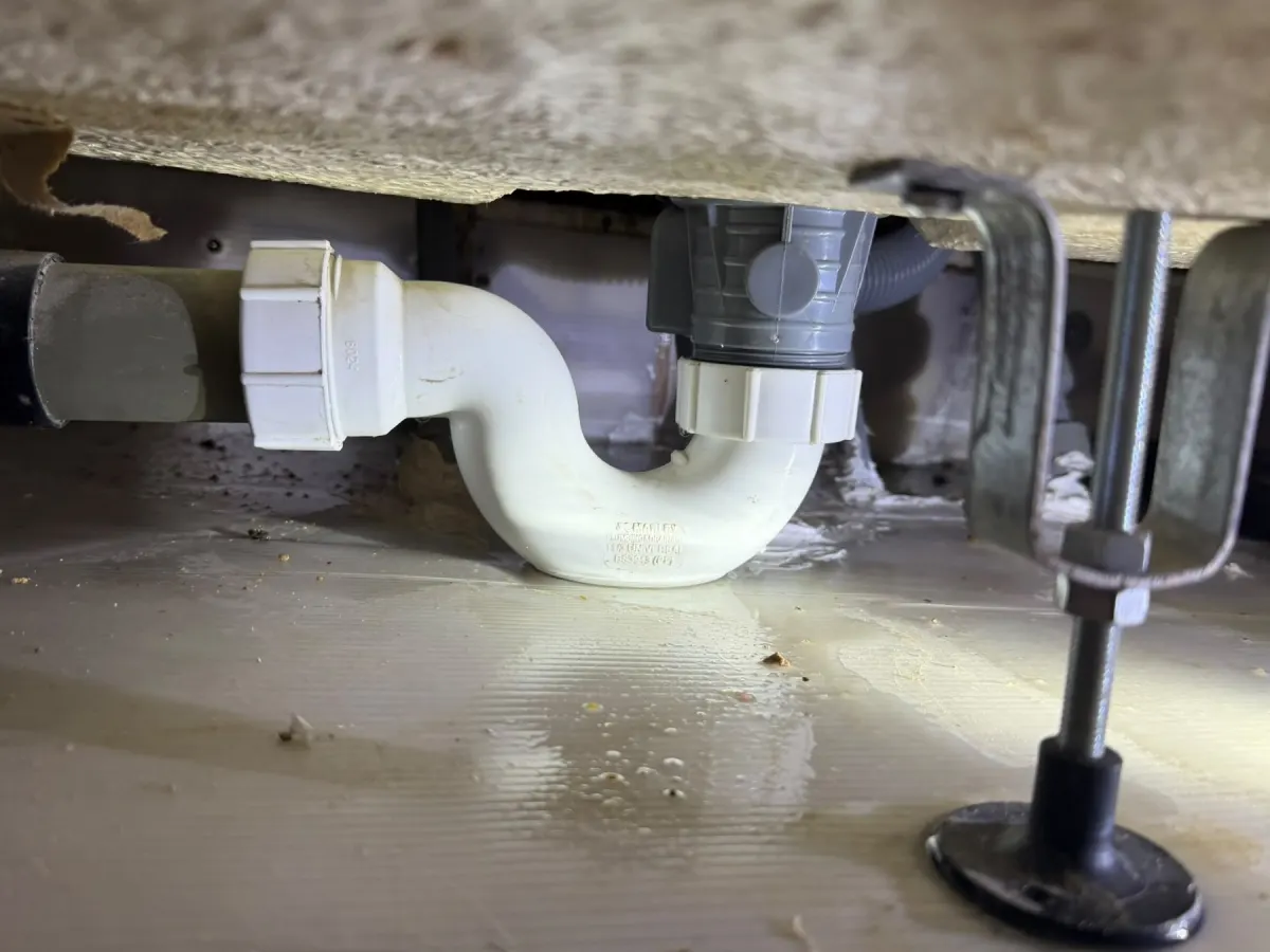 White bath trap and grey waste outlet hanging above a plastic tray with a wet film on it, with a metal clamp at the right