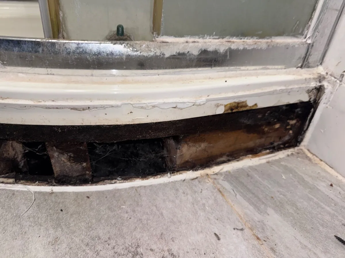 Curved shower tray edge sitting above an open gap, with dark rotted timber blocks and cobwebs visible beneath it
