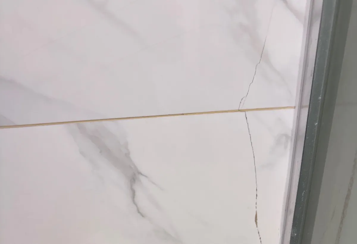 Hairline crack running down through white wall tiles and a grout line beside a glass shower enclosure frame