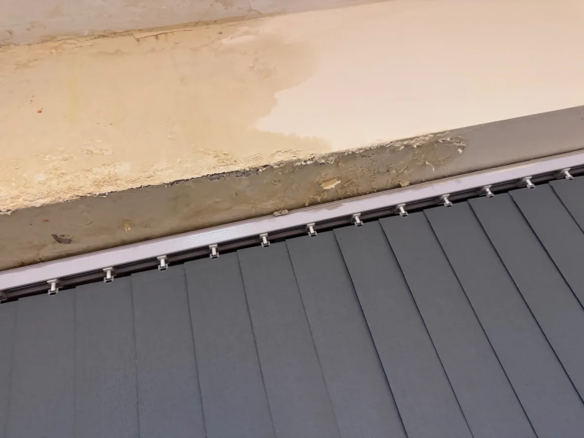 Peeling, stained plaster on the reveal above a grey vertical blind, with a clear tide line where the damp stops