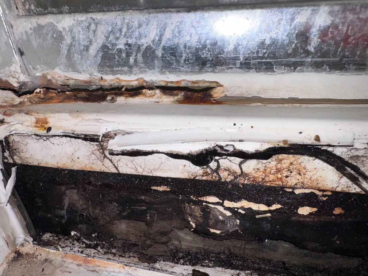 Blackened rotted timber and mould under the edge of a shower tray, with rust staining on the metal frame above it