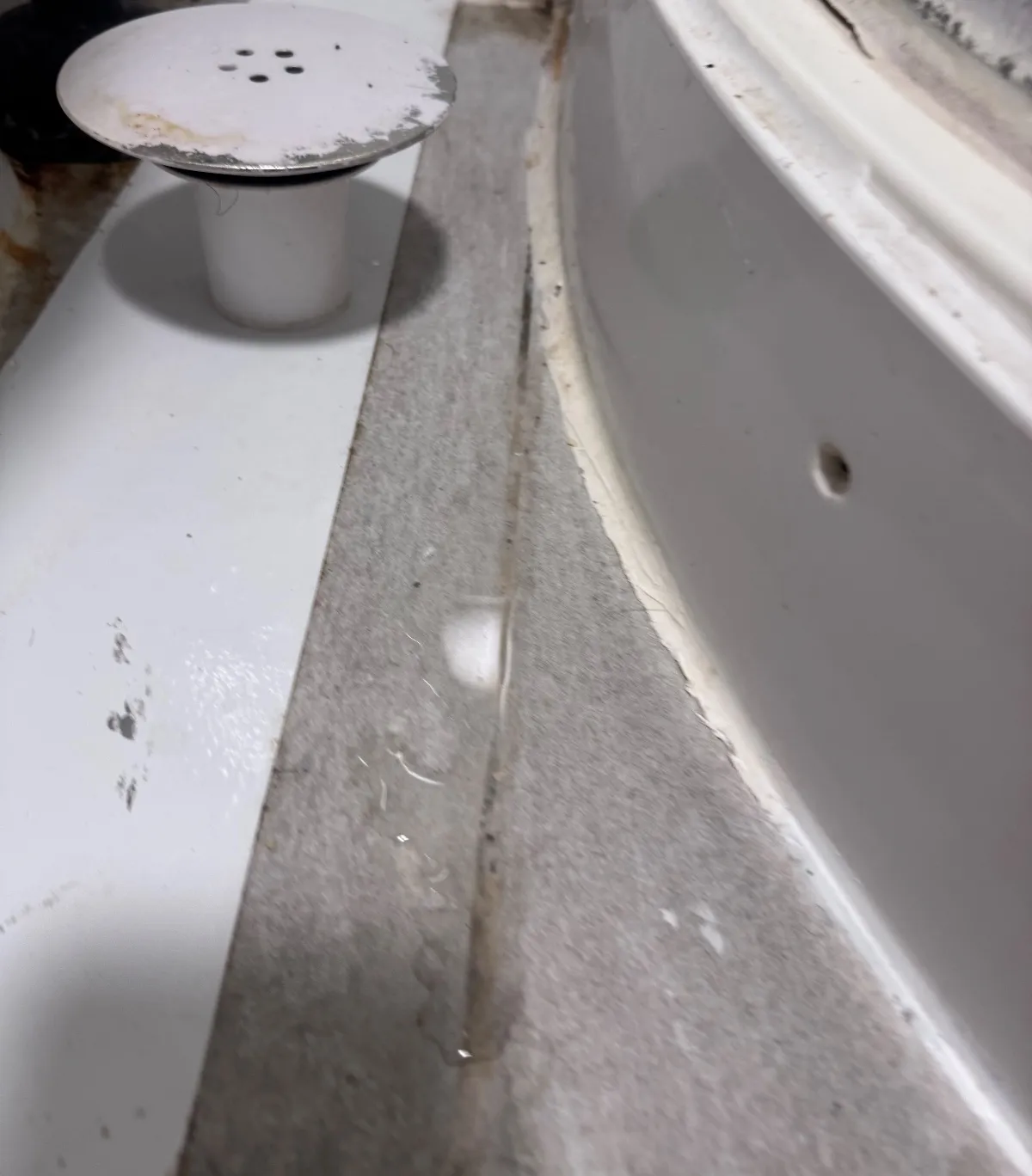 Shower waste cover lifted off its outlet on a floor strip beside a curved tray, with a gap and tile joint beside it