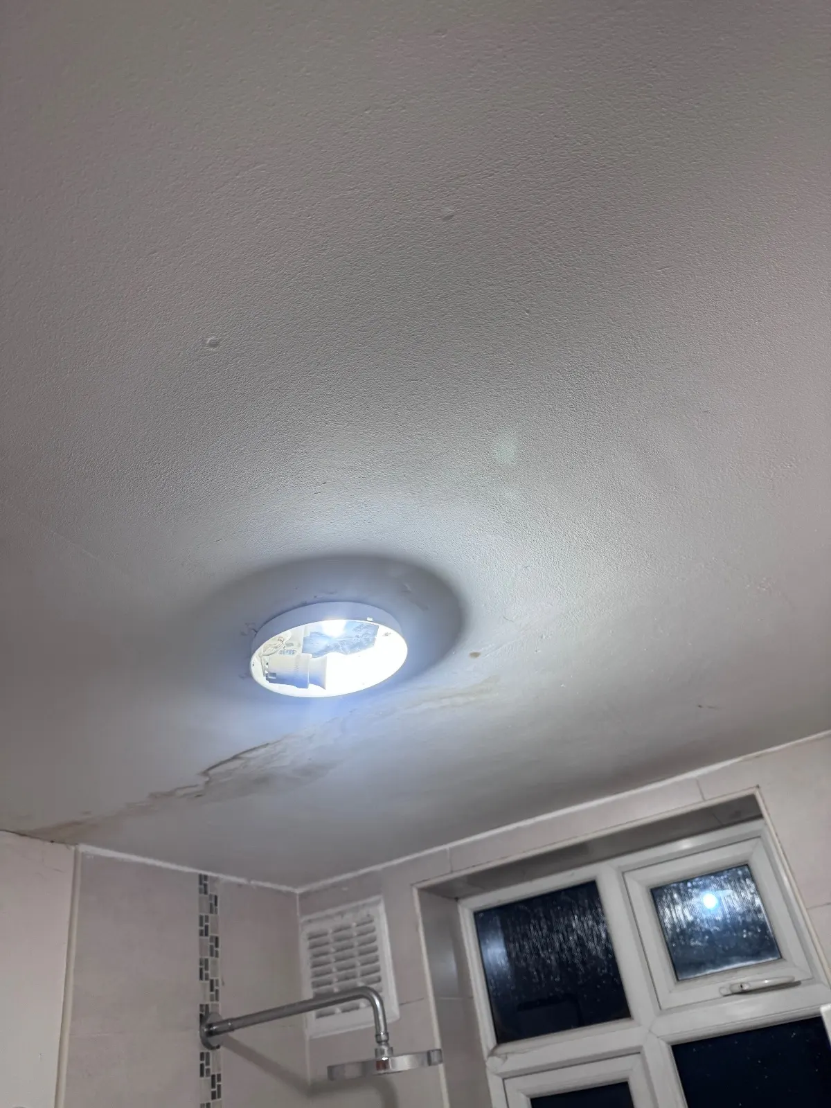Bathroom ceiling with a brown tide mark curving past an open light fitting, with a shower head and window below