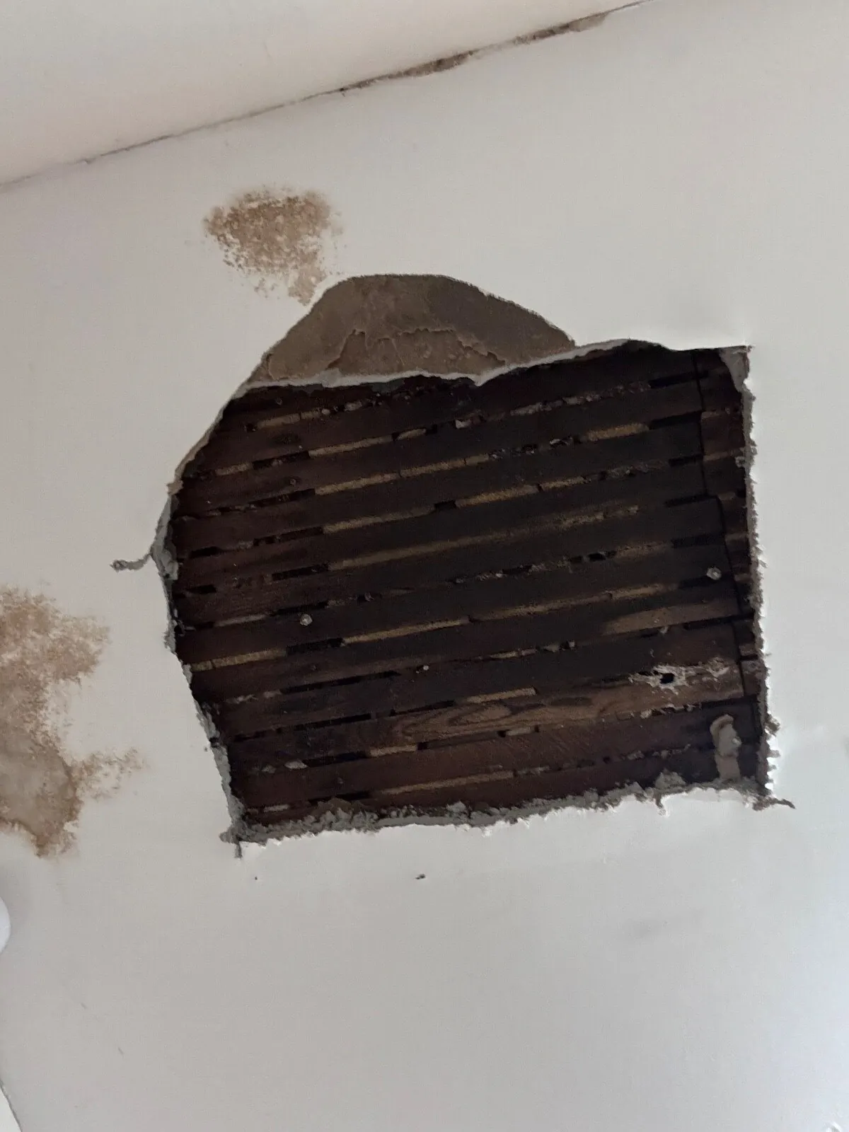 Rectangular opening cut in a ceiling showing dark timber laths, with brown water staining on the paint beside it