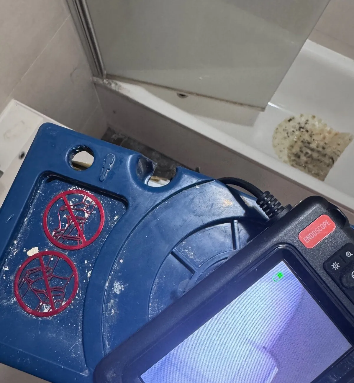 Handheld endoscope monitor held beside a bath with a glass screen, resting on a blue plastic case