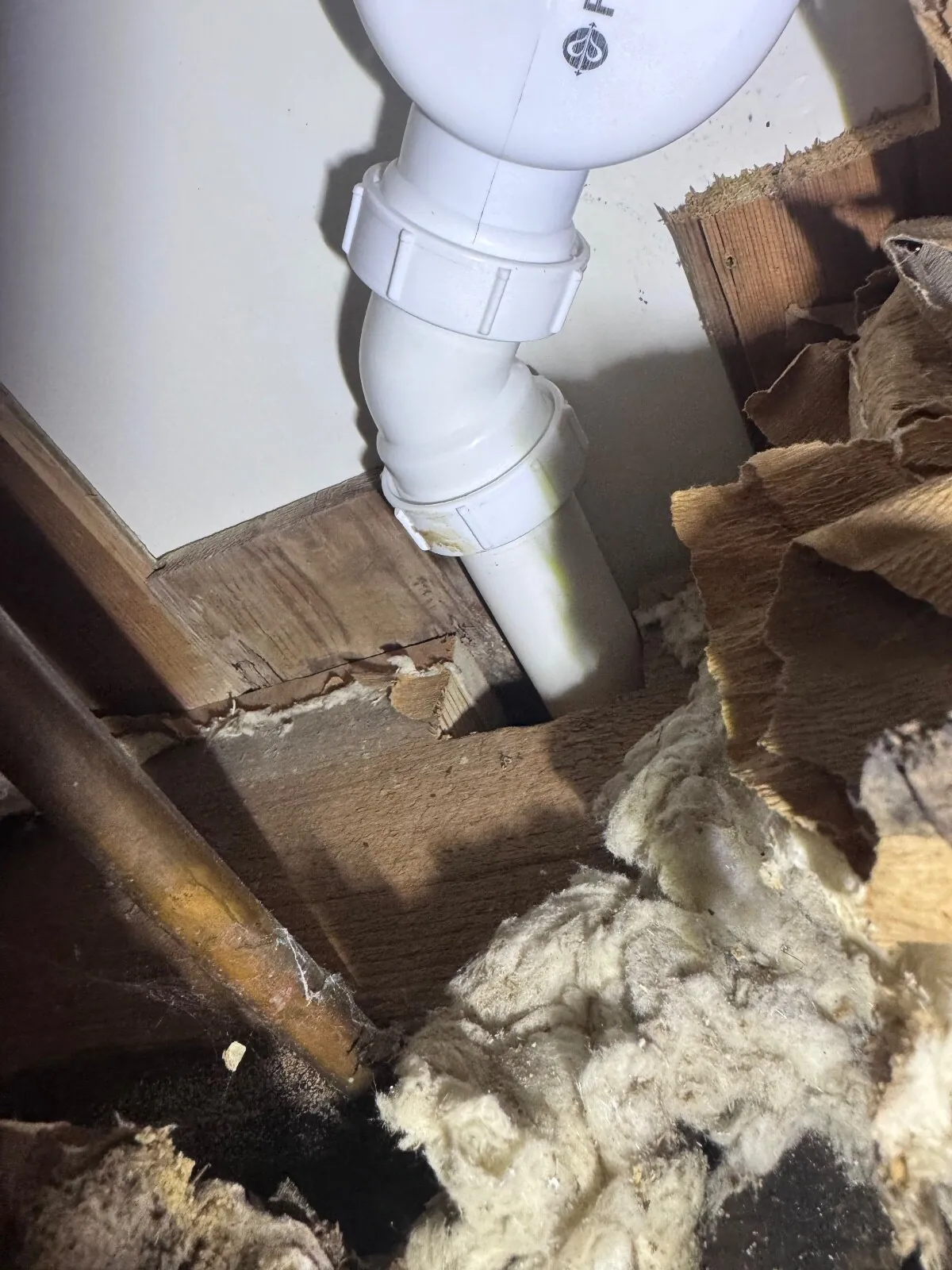 Plastic shower waste trap with two white union nuts in a joist void, loose insulation and a copper pipe beside it