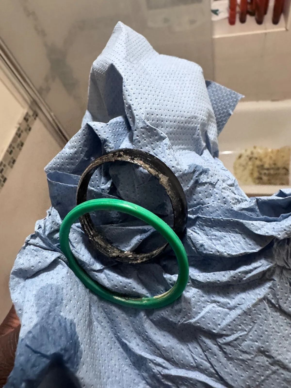 A worn black rubber sealing ring and a green ring lying on a blue paper towel, with a bathroom wall behind