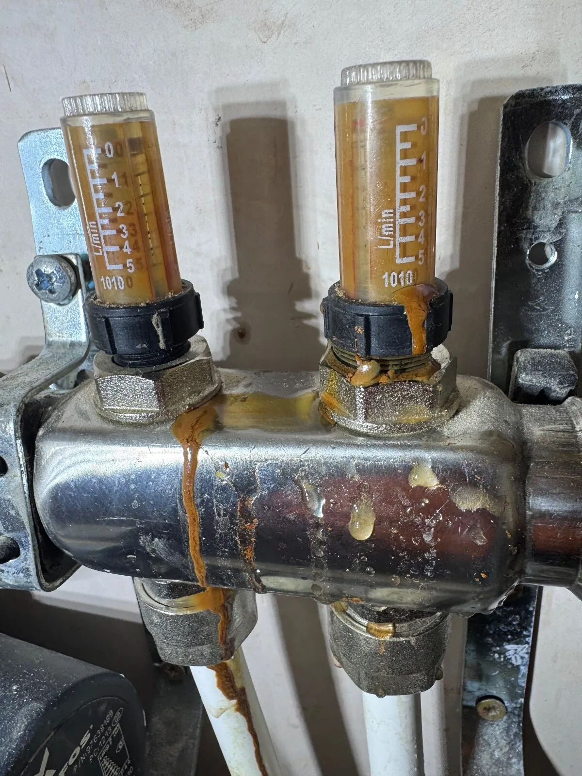 Underfloor heating manifold flow meters with orange rust staining running down the body and water droplets on the metal