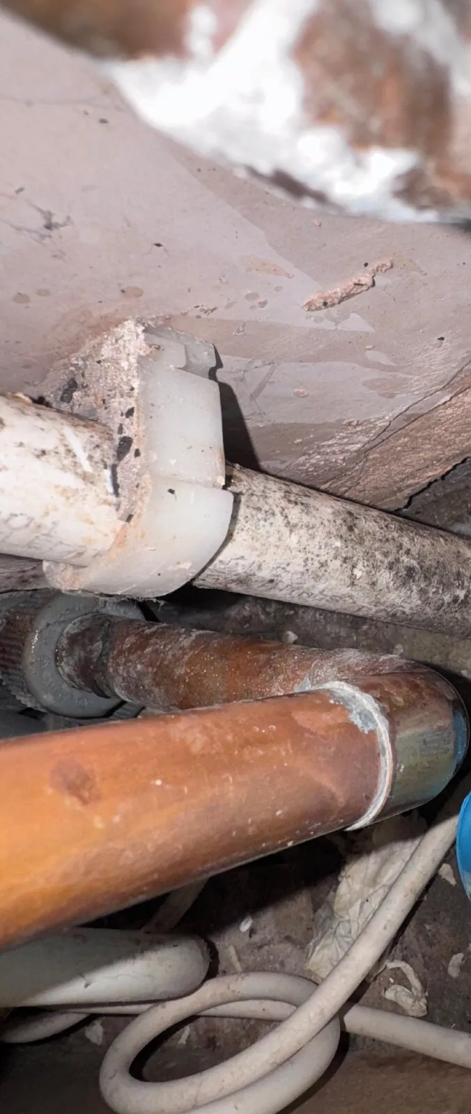 Copper pipe and a clipped plastic pipe in a dusty void beneath a floor, under a rough cracked surface