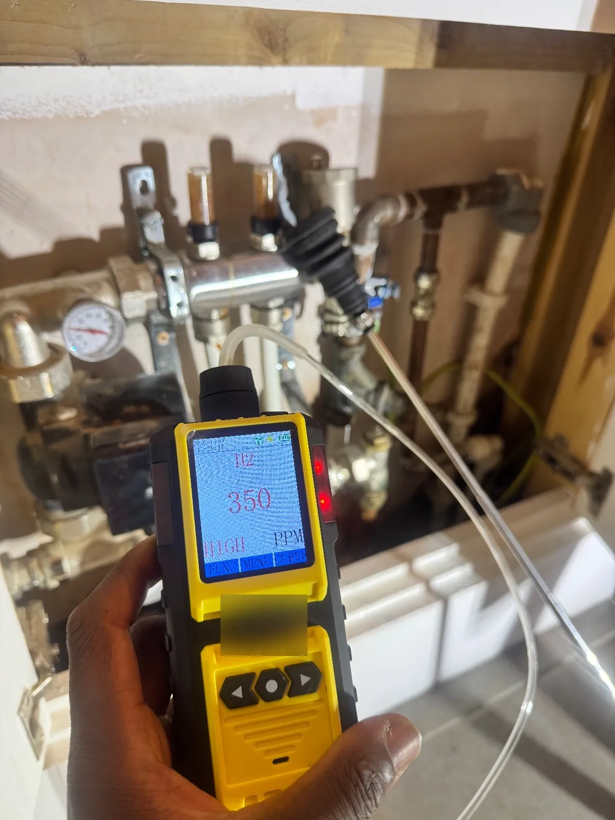 Hand holding a hydrogen detector reading 350 ppm and HIGH, with the sampling tube running to pipework in a manifold cupboard