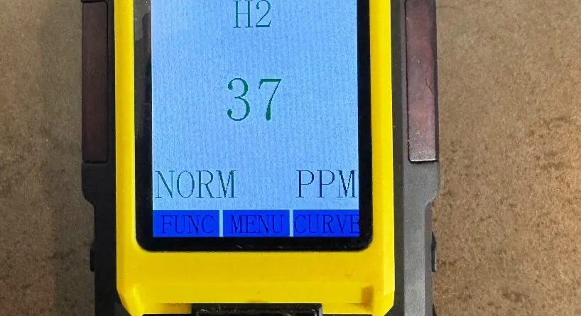 Close view of a hydrogen detector screen headed H2 and reading 37 ppm, marked NORM, in a yellow and black case