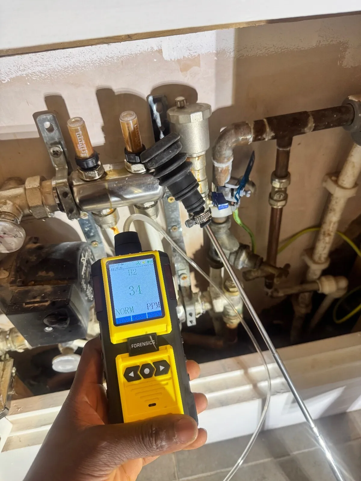 Hydrogen detector showing 34 ppm and a NORM marker, with the probe tip resting at a fitting beside a heating manifold