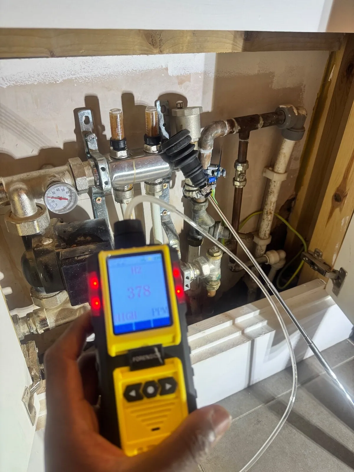 Hydrogen detector showing 378 ppm and a HIGH marker, held in front of a heating manifold with the probe at a fitting