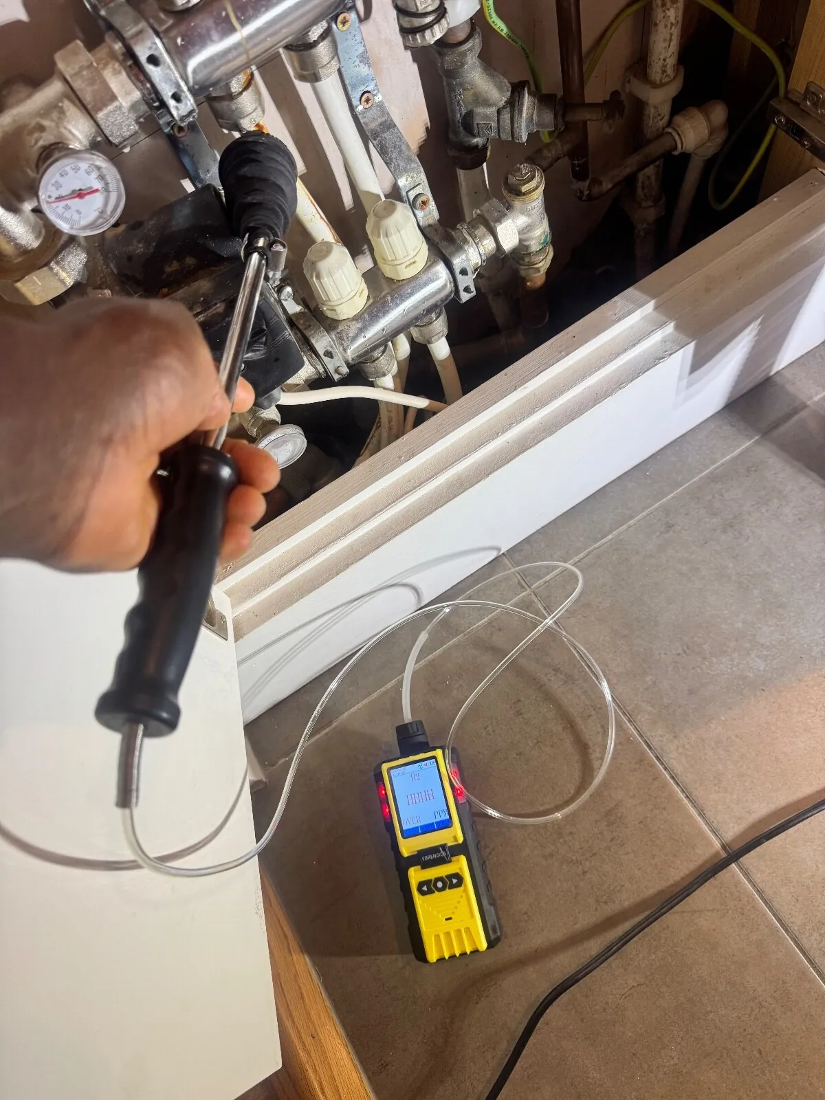 Hand holding a long gas probe towards a heating manifold cupboard, with a yellow hydrogen detector on the tiled floor