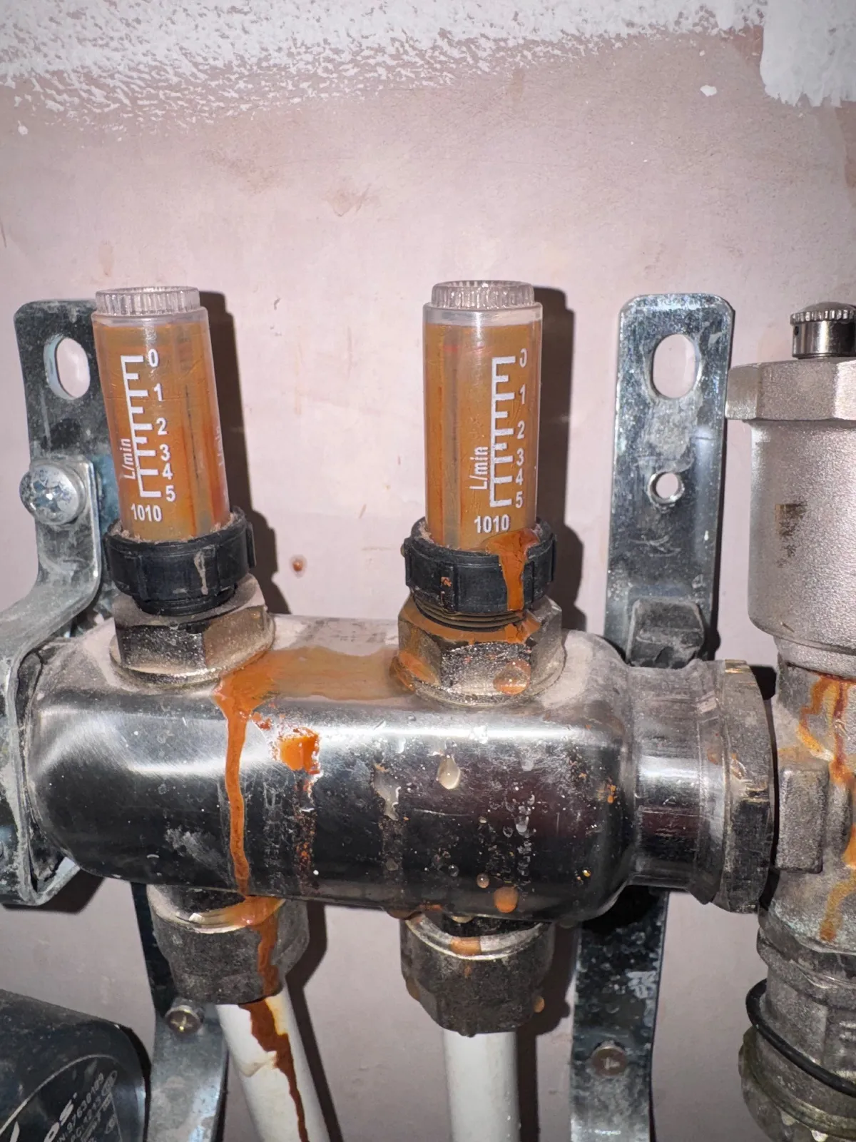 Two flow meter tubes on an underfloor heating manifold with orange staining on the fittings, manifold body and pipes below