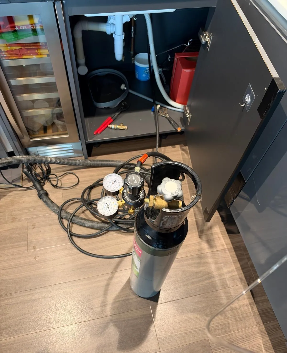 Gas cylinder with a two-gauge regulator and hoses on a wood-effect floor in front of an open kitchen sink cupboard