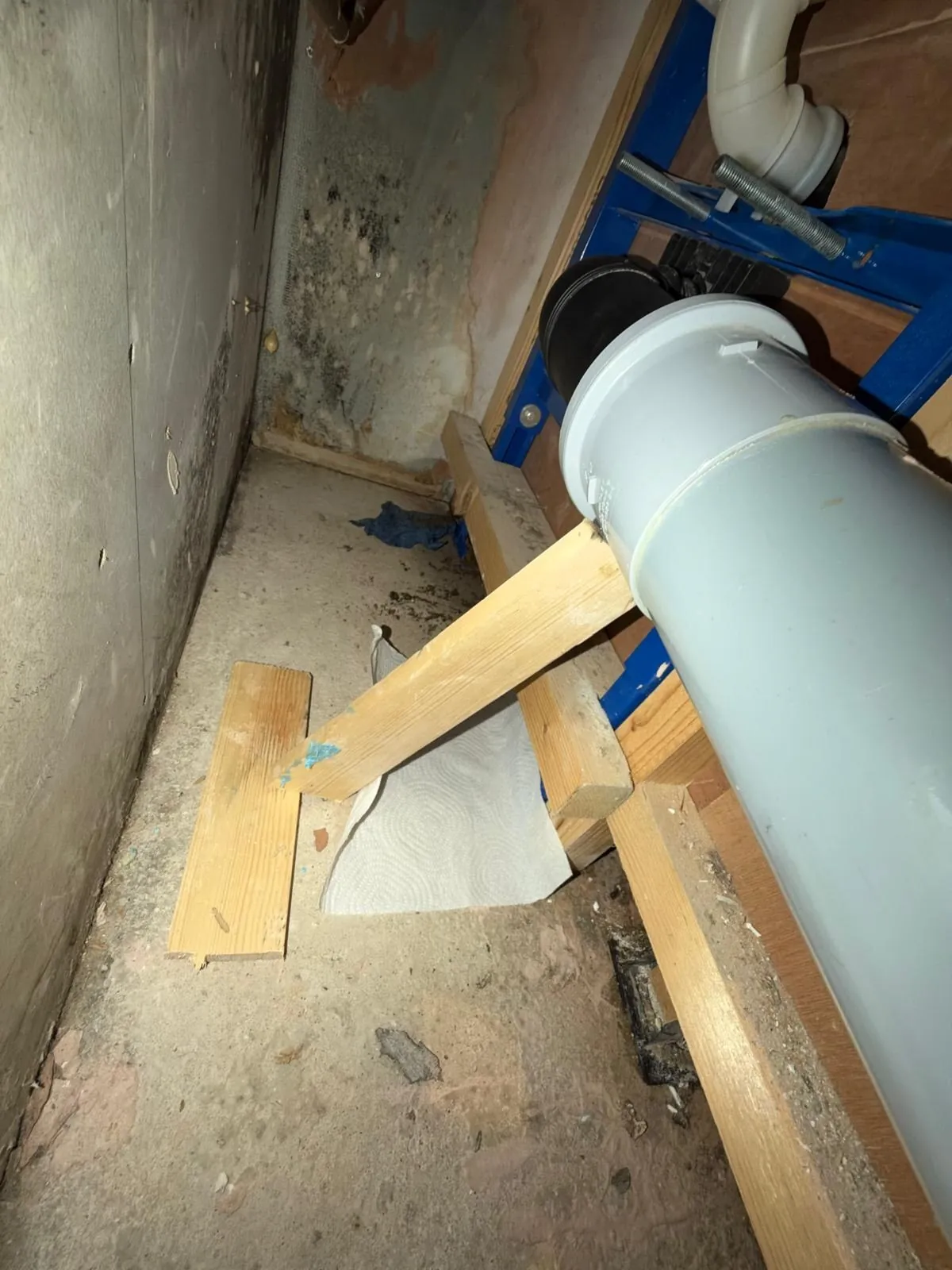Grey soil pipe running through a duct beside a blue toilet frame, resting on timber, with dark mould on the wall behind