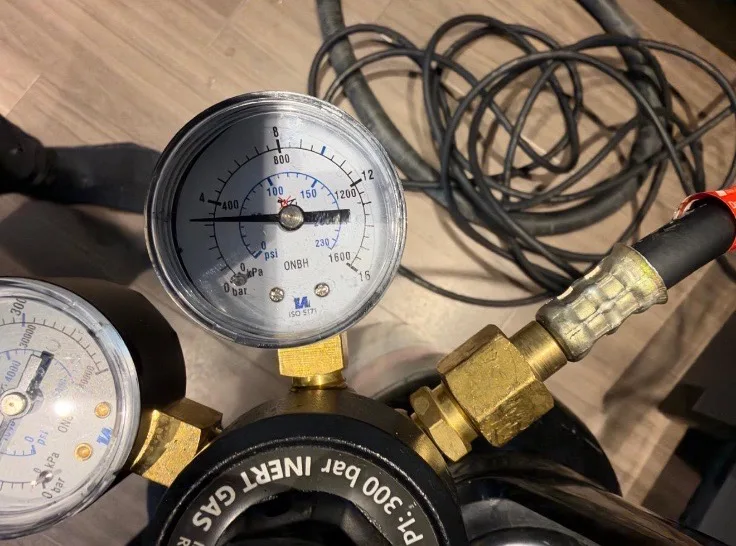 Inert gas regulator with two pressure gauges and a braided hose, resting on a wood-effect floor beside coiled cable