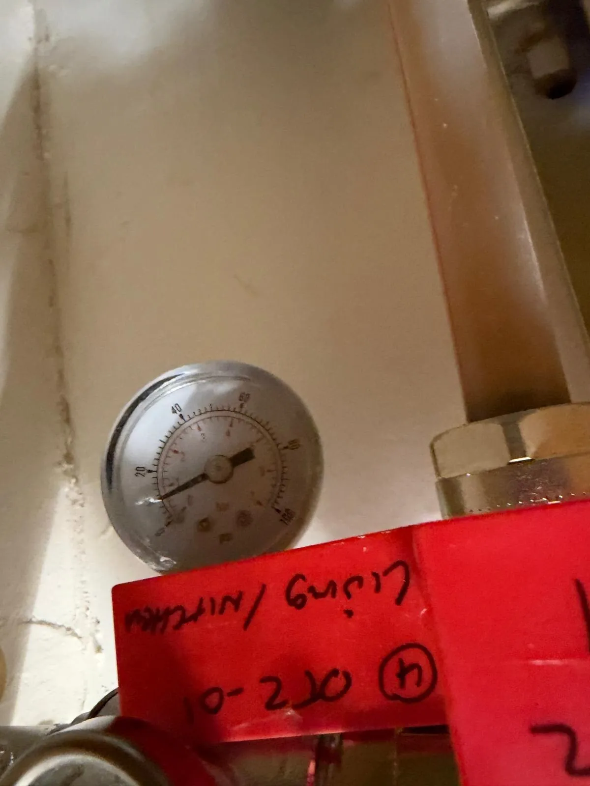 Round pressure gauge with a 0 to 100 dial, mounted above a red tag carrying handwritten labels