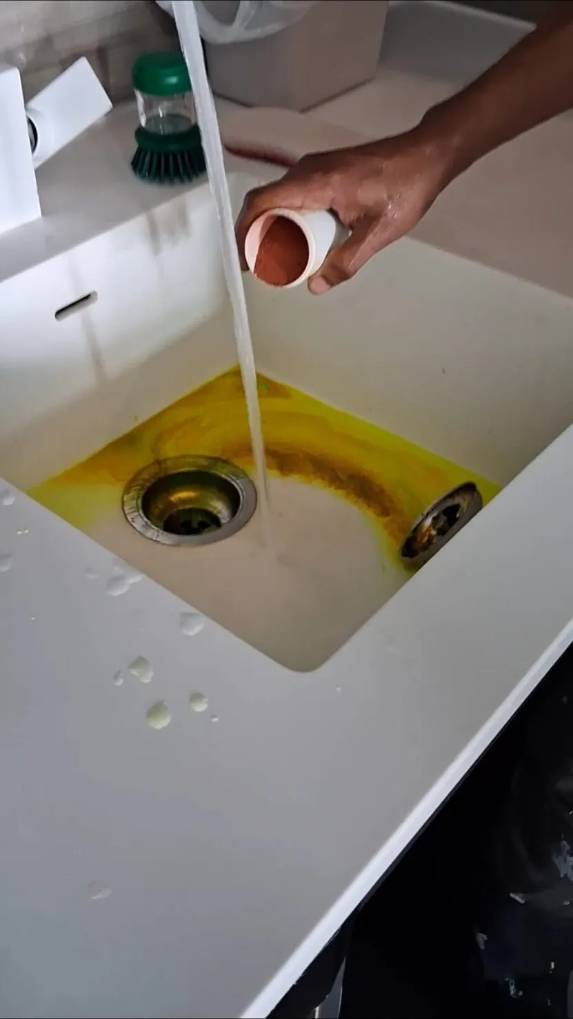 Hand pouring orange powder into a running tap over a white sink, with a yellow dye trail curling round the plughole
